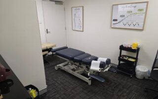 Chiropractic treatment room at Northbridge Chiropractic in Perth with a navy blue adjustable treatment table, anatomical spine poster, and wellness diagram on the wall.