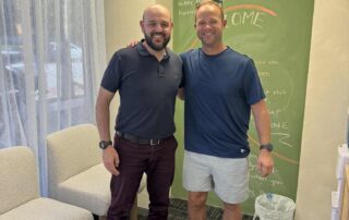 Dr Mario Vinci chiropractor at Northbridge Chiropractic in Perth smiling with a patient in the clinic waiting area in front of the green chalkboard feature wall.