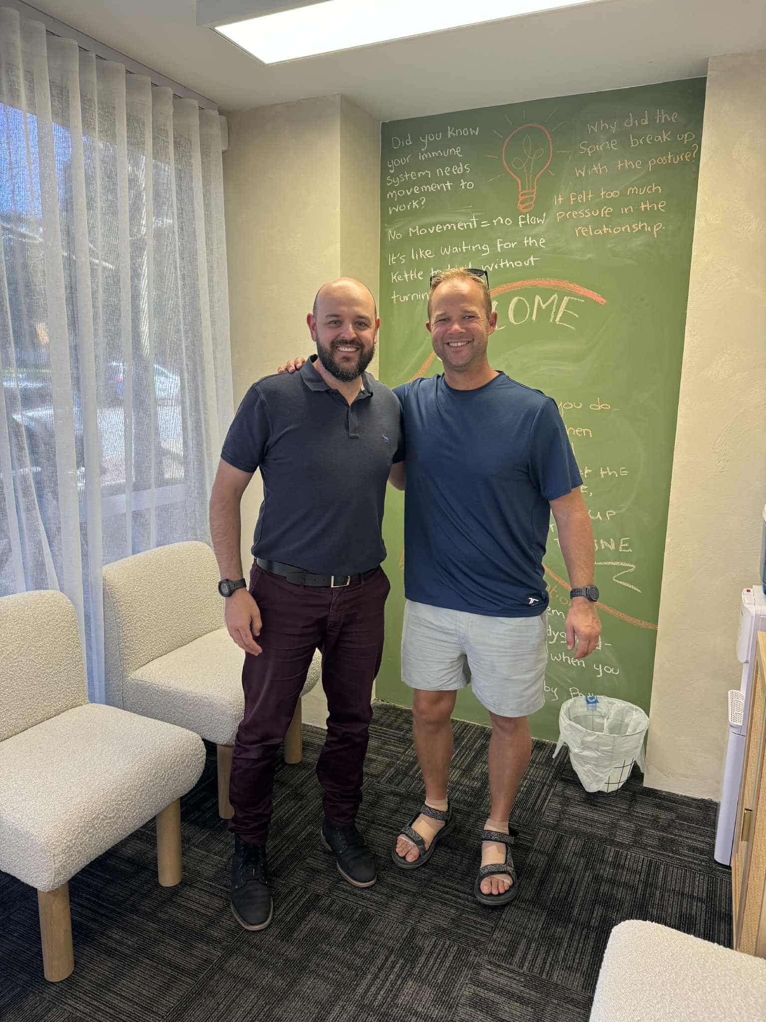 Dr Mario Vinci chiropractor at Northbridge Chiropractic in Perth smiling with a patient in the clinic waiting area in front of the green chalkboard feature wall.
