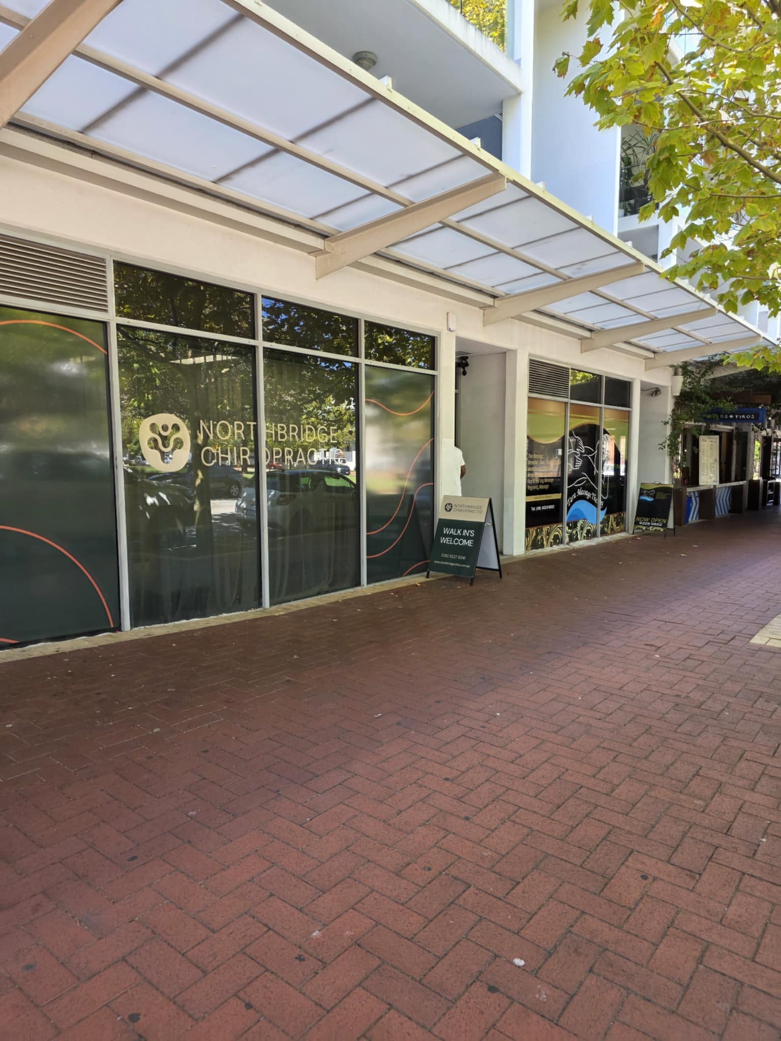 Angled street view of Northbridge Chiropractic clinic frontage in Perth showing the clinic logo on the shopfront window and a walk-ins welcome sign on the footpath beneath a covered arcade.