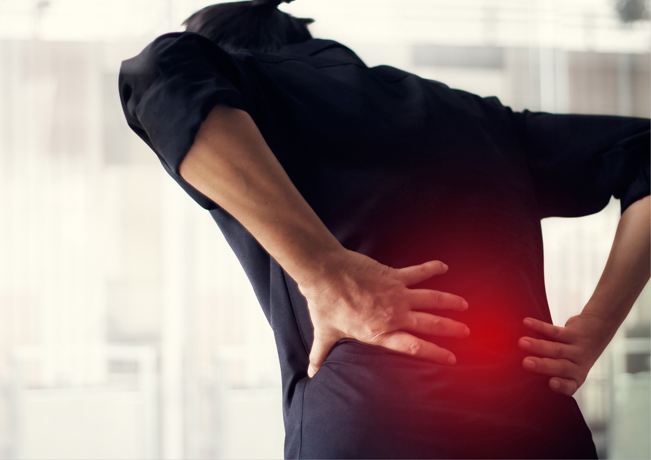 Person in a dark top bending slightly forward with both hands pressed against her lower back, with a red highlighted area indicating lumbar spine discomfort in an office environment.