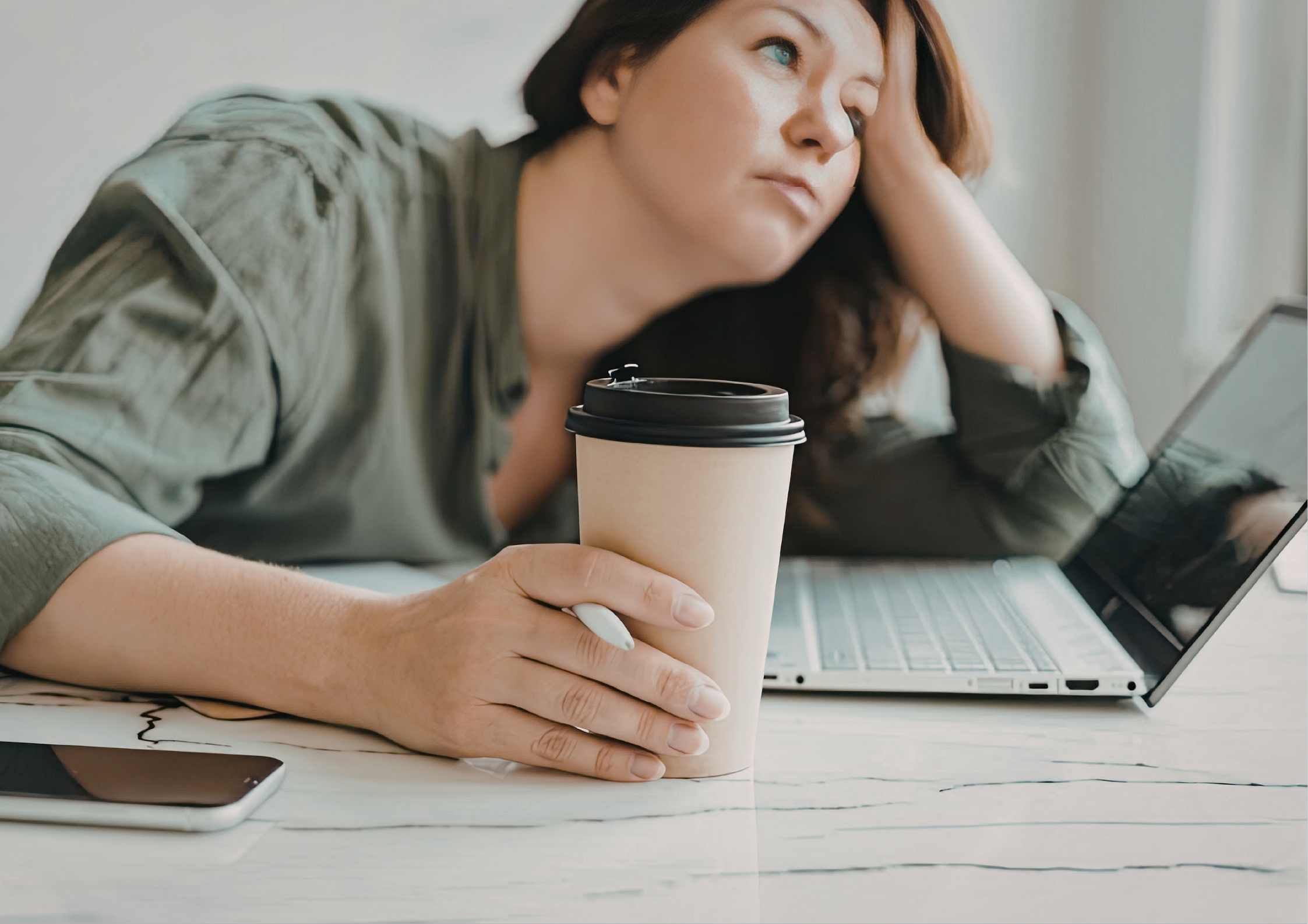 Woman leaning on a desk with her head resting on one hand and holding a takeaway coffee cup, looking upward with a distracted and fatigued expression, illustrating mental fatigue, burnout, and physical exhaustion from prolonged desk work.