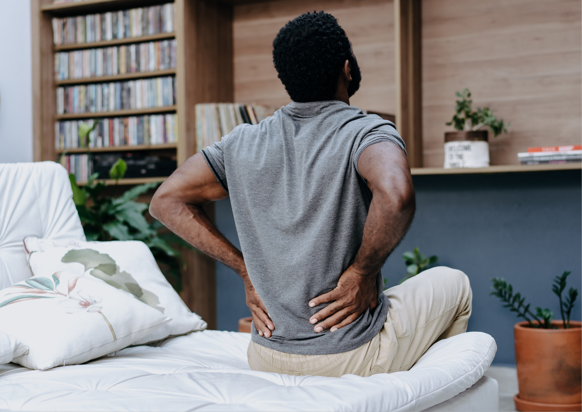 Man sitting on the edge of a bed with both hands pressed against his lower back, illustrating lumbar spine discomfort and lower back pain in an everyday home setting.