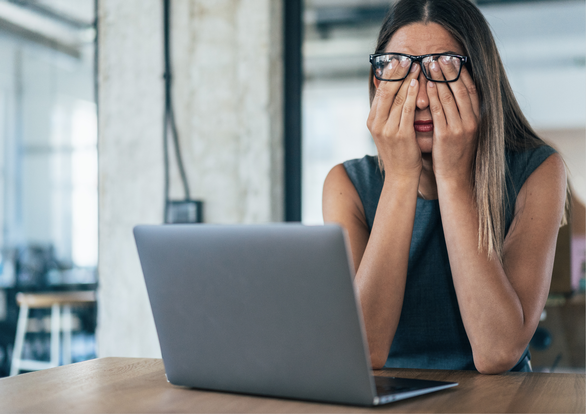 Woman sitting at a laptop in an office pushing her glasses up and covering her eyes with both hands, illustrating workplace stress, eye strain, and tension commonly associated with desk work and prolonged screen time.