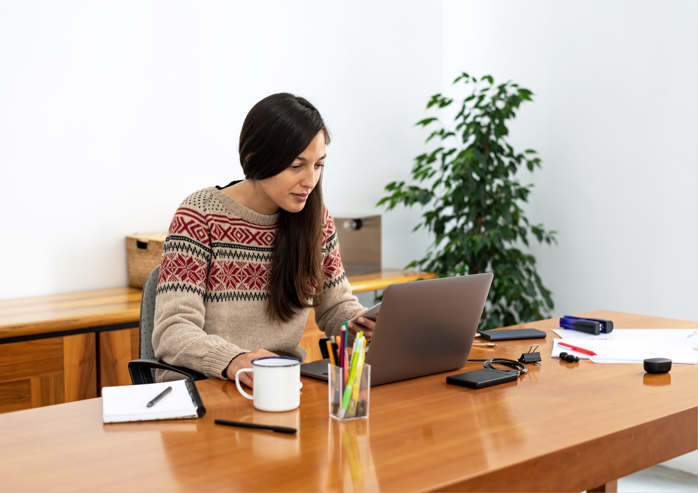 Woman with long dark hair wearing a patterned knit jumper sitting at a timber home office desk leaning forward toward a laptop, illustrating forward head posture and rounded shoulder position during desk work and remote working.