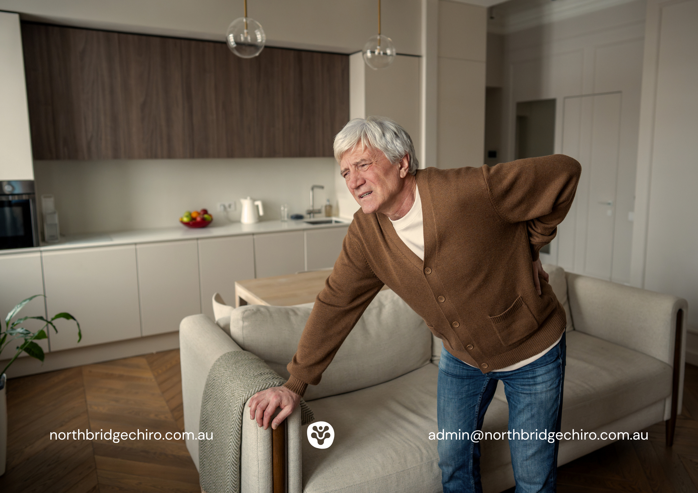 Older man with white hair wearing a brown cardigan leaning on the back of a sofa and holding his lower back with a pained expression, illustrating lower back pain and lumbar discomfort in an everyday home setting.