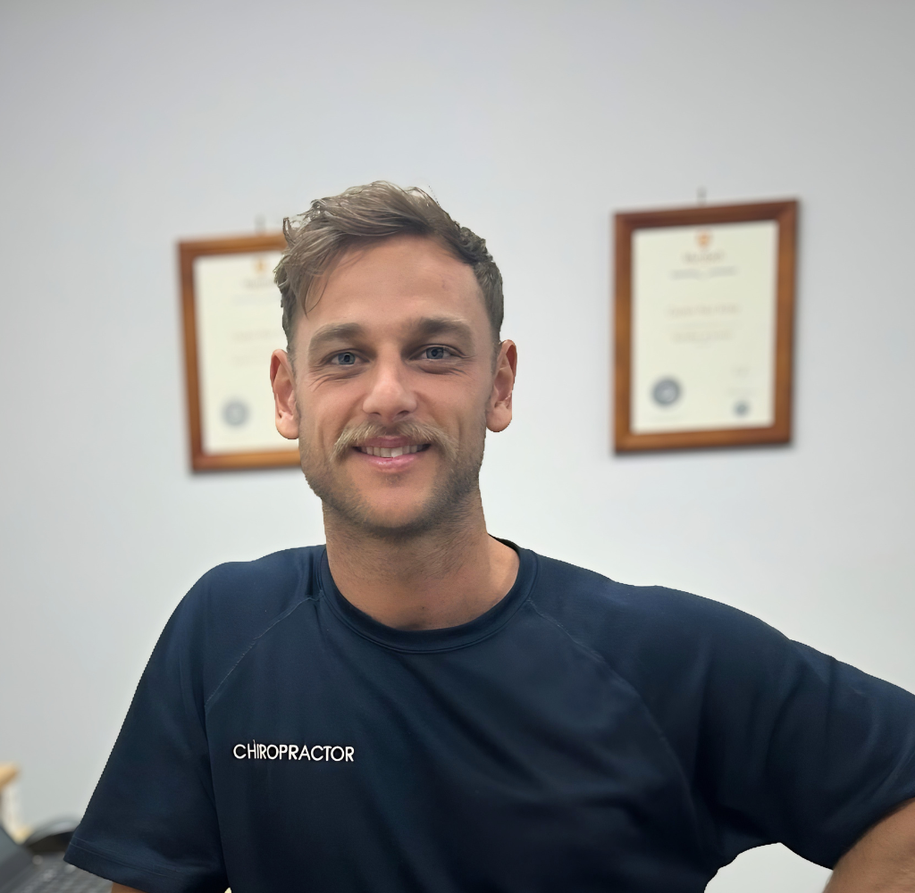 Dr Clay chiropractor at Northbridge Chiropractic in Perth smiling in the clinic with two framed professional qualifications displayed on the wall behind him.